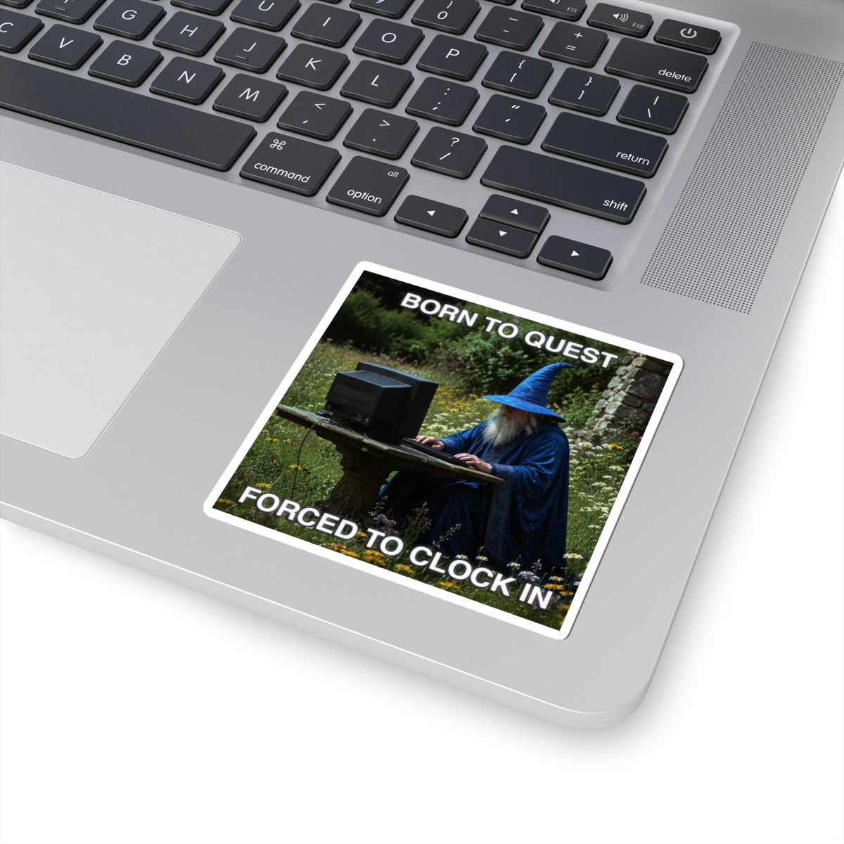 Laptop with a sticker of a wizard using a computer and text 'Born to quest, forced to clock in' on a grassy background.