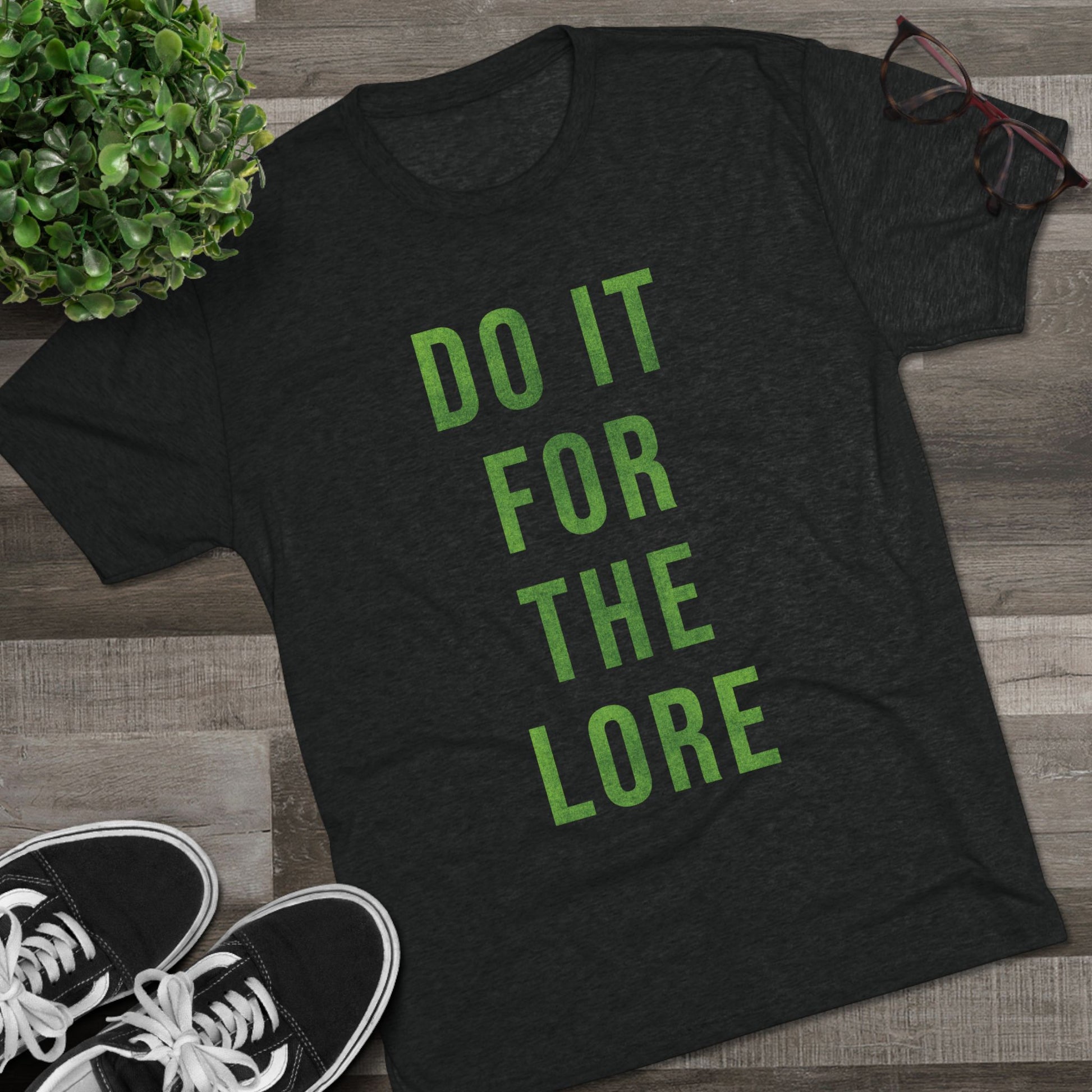 Black t-shirt with 'DO IT FOR THE LORE' text on a wooden surface with sneakers and a plant.