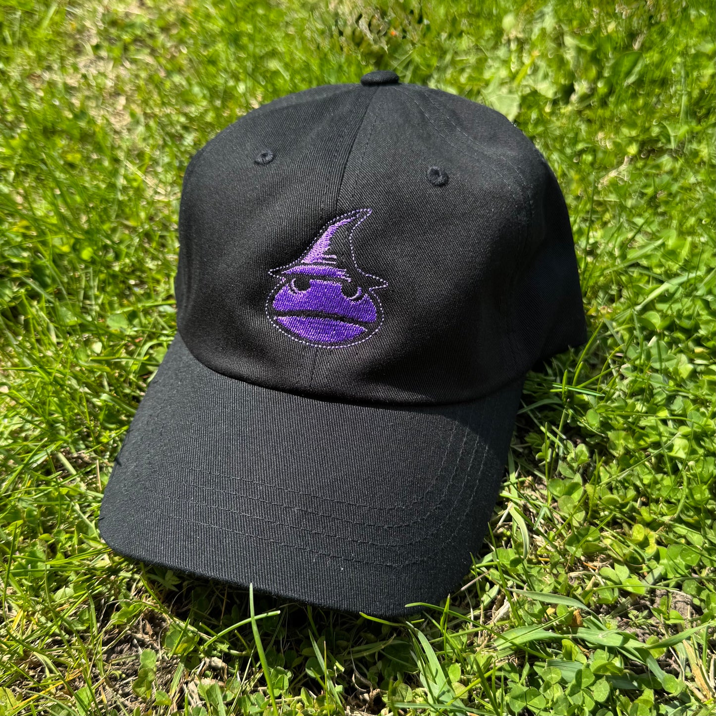A Classic Black Dad Hat with a purple frog wizard logo on the front. The hat is laying in the grass