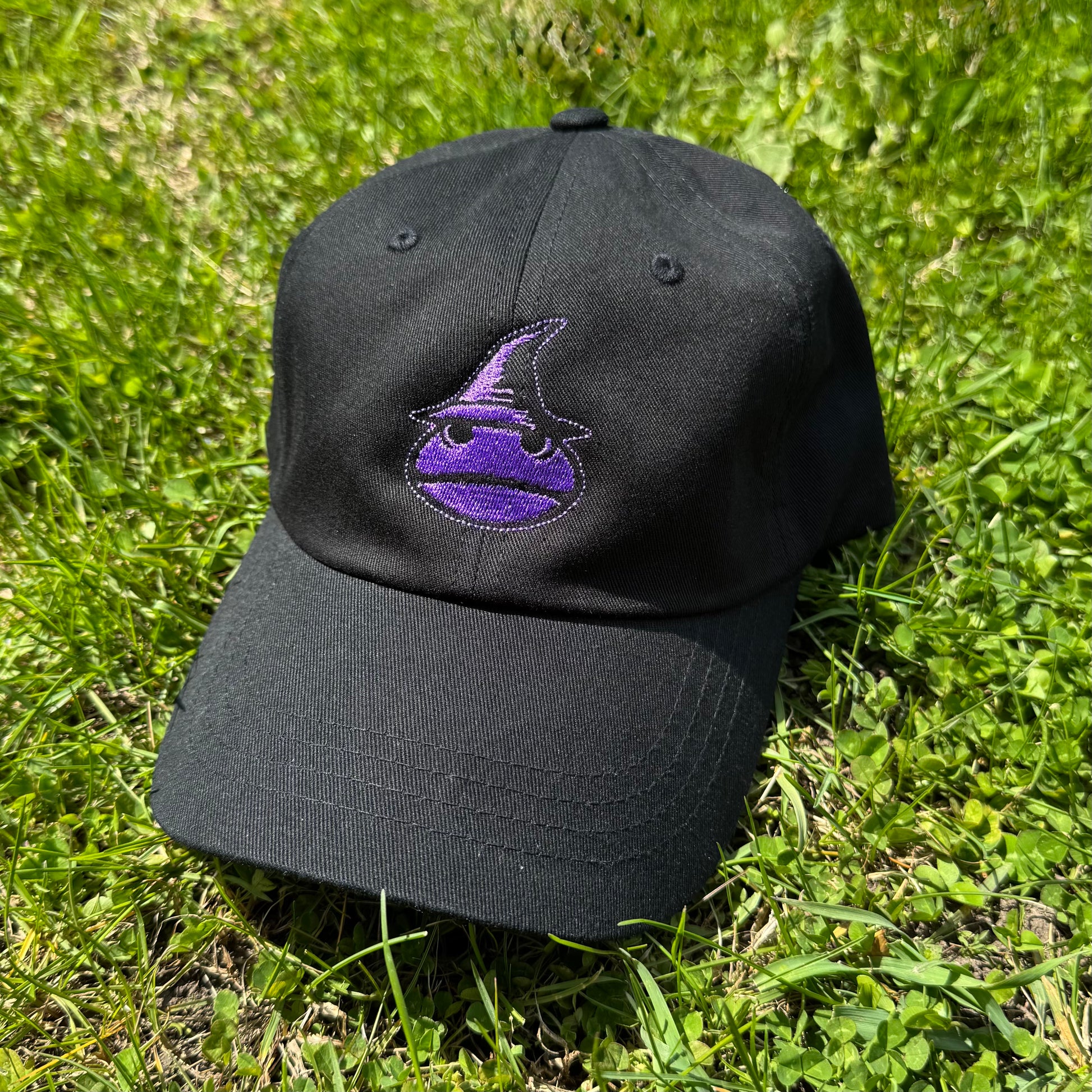 A Classic Black Dad Hat with a purple frog wizard logo on the front. The hat is laying in the grass
