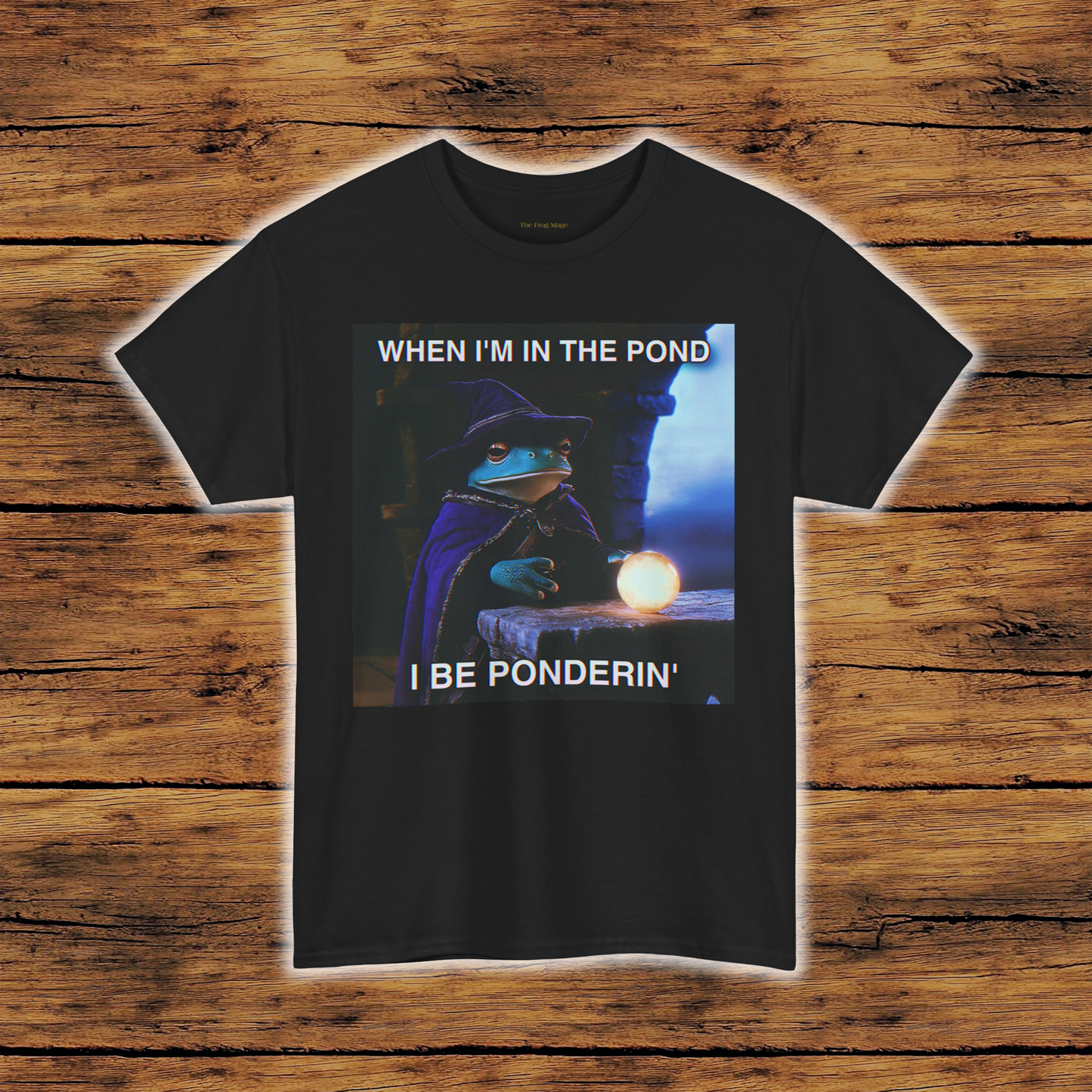 Black T-Shirt with a graphic of the frog mage sitting at a table with a glowing orb on it in front of him with text that reads "WHEN I'M IN THE POND I BE PONDERIN'" in a dark castle setting